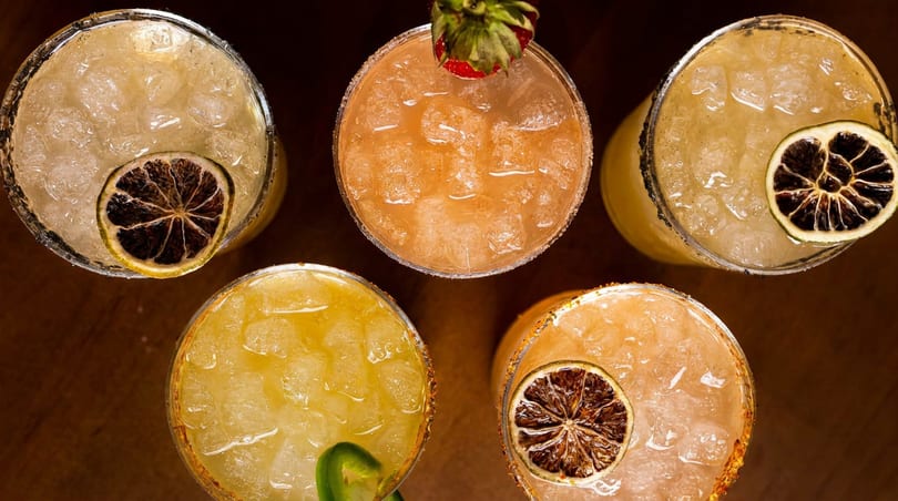 Overhead view of five colorful margaritas with ice, garnished with dried citrus, strawberry, and jalapeño.