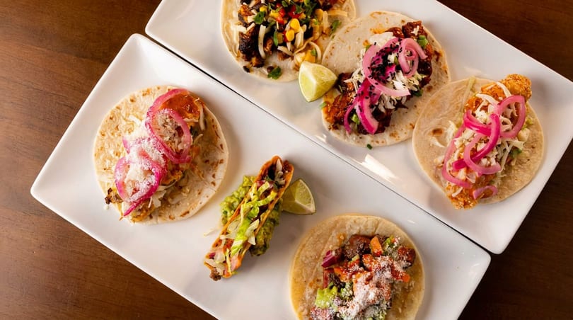 Two white rectangular plates filled with various tacos, including one hard-shell taco with guacamole.