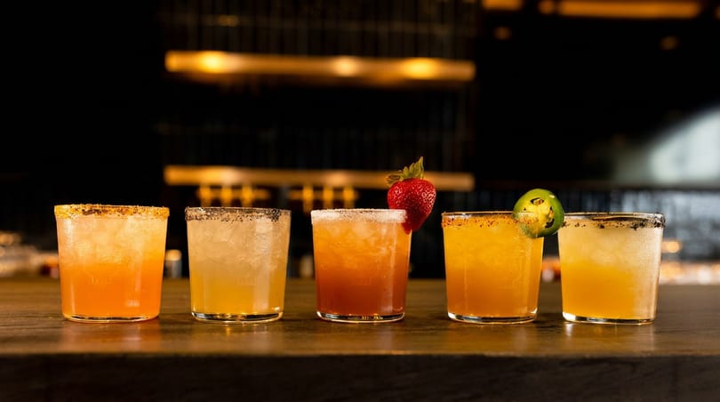 Five colorful cocktails with various garnishes, including strawberry and jalapeño, on a dark bar.