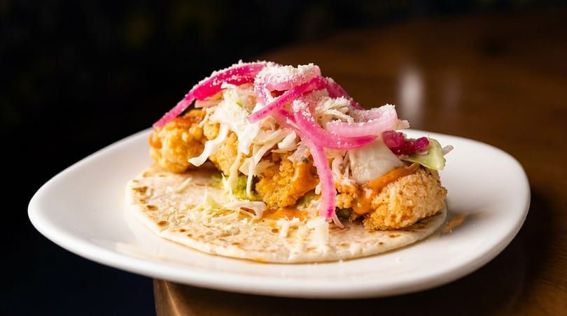 Crispy fried chicken taco with pickled red onions, shredded cabbage, and cotija cheese on a white plate.