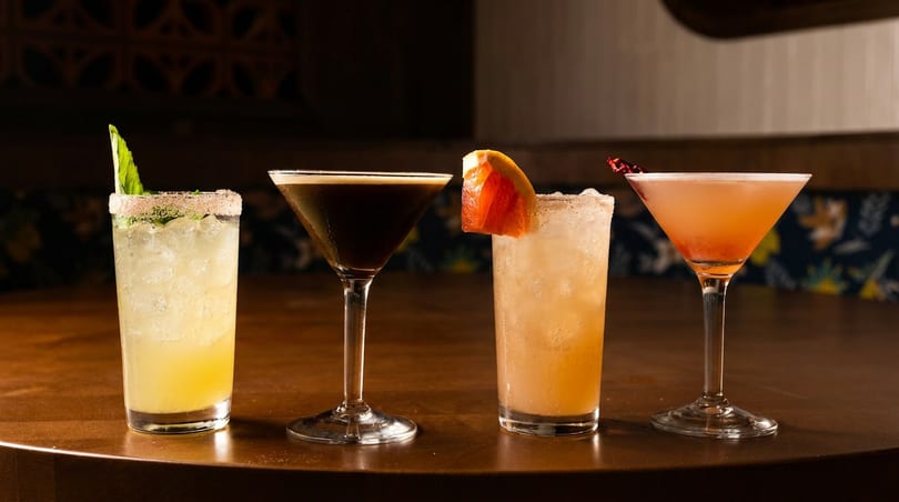 Four colorful cocktails, including an espresso martini and a grapefruit cocktail, sit on a dark wooden bar.