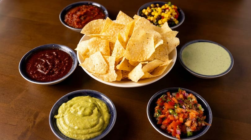 A bowl of tortilla chips surrounded by six bowls of various salsas and dips on a wooden table.